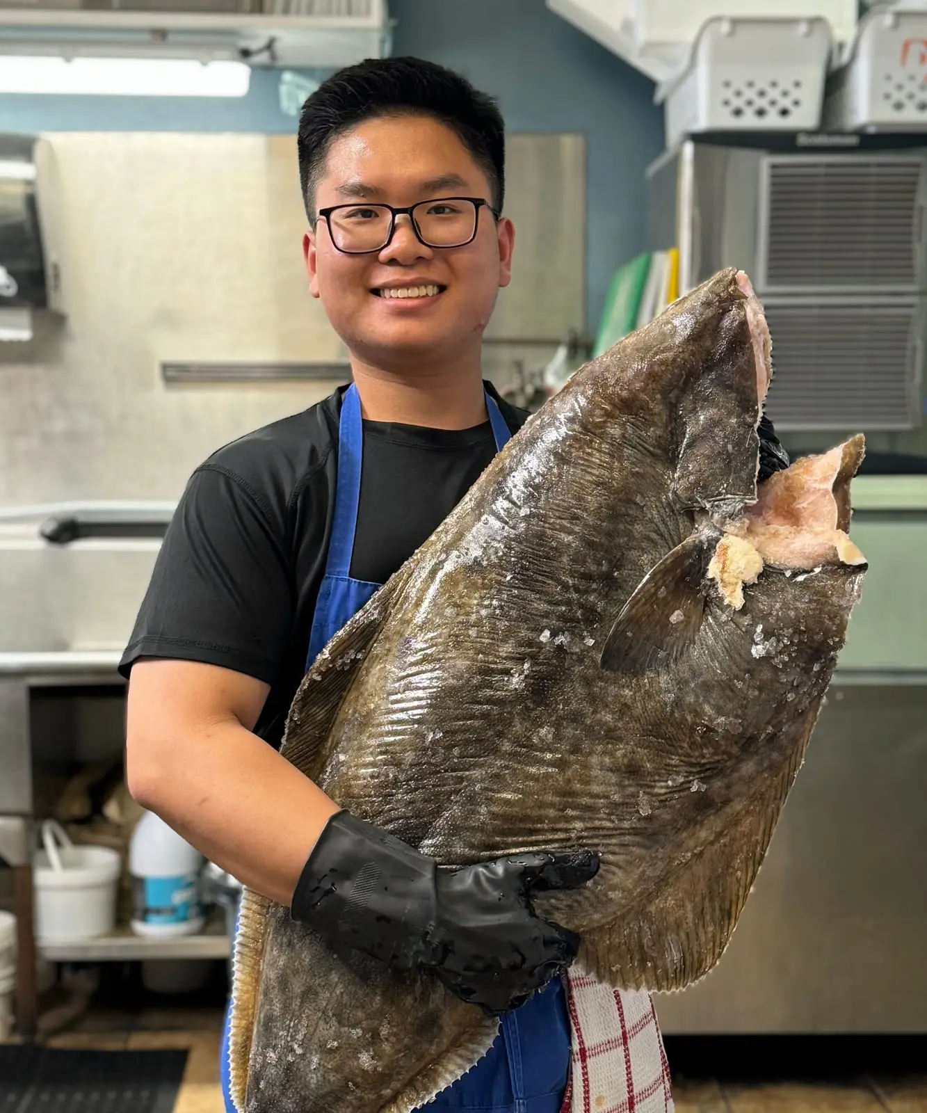 A fishmonger in a blue apron holding a whole Pacific halibut nearly as tall as he is