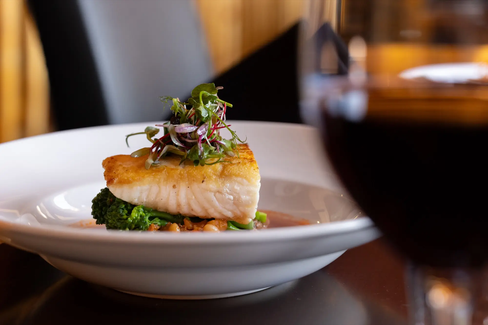 A seared halibut portion plated with microgreens beside a glass of red wine in a dining room
